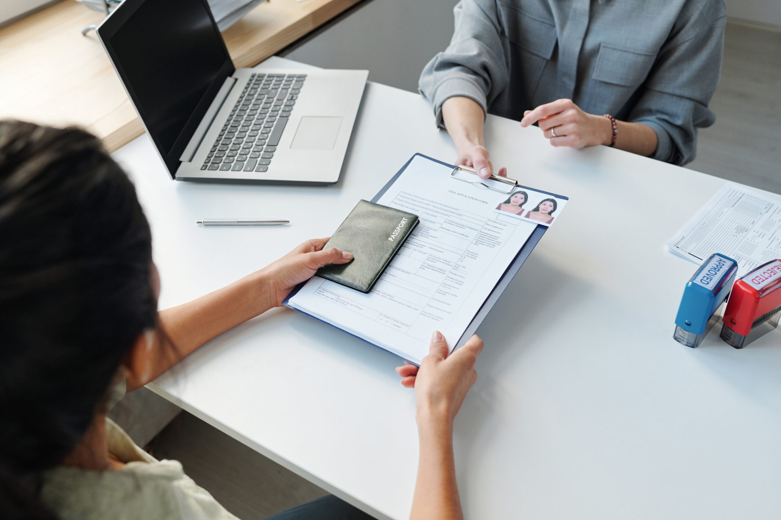 A person sitting at a desk hands over a clipboard with documents and a passport to another person across the table, possibly during an application or verification process. A laptop, pen, and office stamps are visible on the desk.