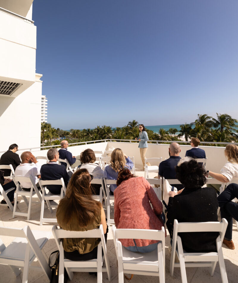 Conference attendees seated for an outdoor presentation on a sunny terrace overlooking palm trees and the ocean.